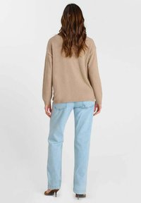 Femme aux longs cheveux ondulés portant un pull beige, un jean bleu clair et des talons marron, debout de dos sur un fond uni.