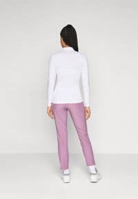 Žena s pletenicama koja nosi bijelu dugu majicu, mauve hlače, bijele čarape i bijele tenisice, stoji pred jednostavnom pozadinom.