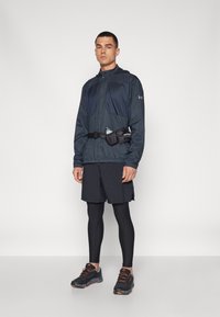 Veste légère bleu foncé avec fermeture éclair, associée à un short noir et des leggings noirs ajustés. Comprend une ceinture avec porte-bouteille et des baskets noires.