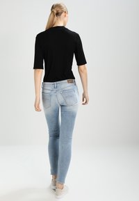 Kobieta z blond końskiem ogonem, ubrana w czarną bluzkę, jasnoniebieskie dżinsy skinny i beżowe sneakersy, idąca w kierunku przeciwnym na jednolitym jasnym tle.