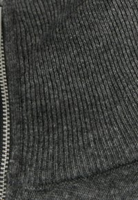 Grijze textuurstof met zichtbare ribbelpatronen en gladde delen, voorzien van een zilveren ritsdetail langs één rand.