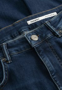 Donkerblauwe spijkerbroek met een gladde textuur, zichtbare contrasterende stiksels, metalen knoopsluiting en een label met de tekst "GABBA" op de tailleband.