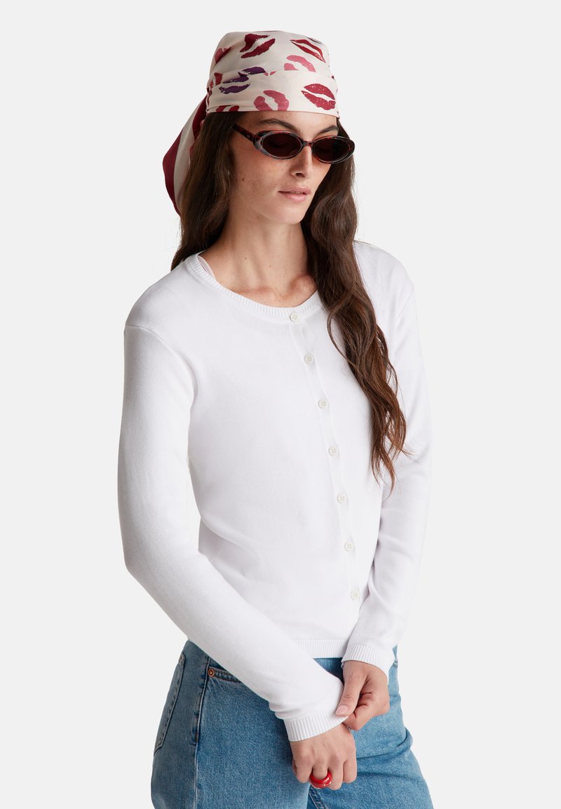 Bílý dlouhý svetr s dlouhým rukávem, kulatým výstřihem, zapínáním na knoflíky a žebrovanými manžetami, spárovaný s modrými džíny a vzorovaným šátkem.