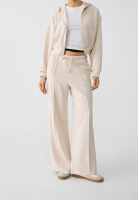 Sudadera corta beige y pantalones de pierna ancha a juego, hechos de una tela suave y lisa. Los pantalones tienen una cintura con cordón; ambos presentan costuras mínimas.