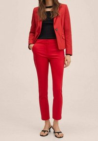 Blazer rouge ajusté avec une fermeture à un bouton, associé à un pantalon slim rouge assorti. Haut noir en dessous, et sandales noires type tongs.