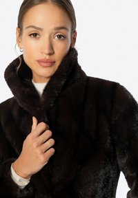 Zwarte nep bonten jas met een hoge kraag, een gladde textuur en een zachte glans. Het model houdt de kraag dicht bij haar hals om de pluche stof te tonen.