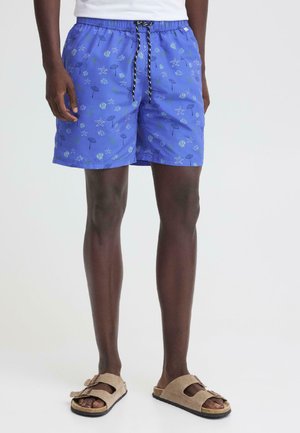 Mężczyzna w fioletowych szortach kąpielowych z wzorem muszelek i rozgwiazd, czarnym sznurkiem, beżowych klapkach, stoi na tle białego tła.