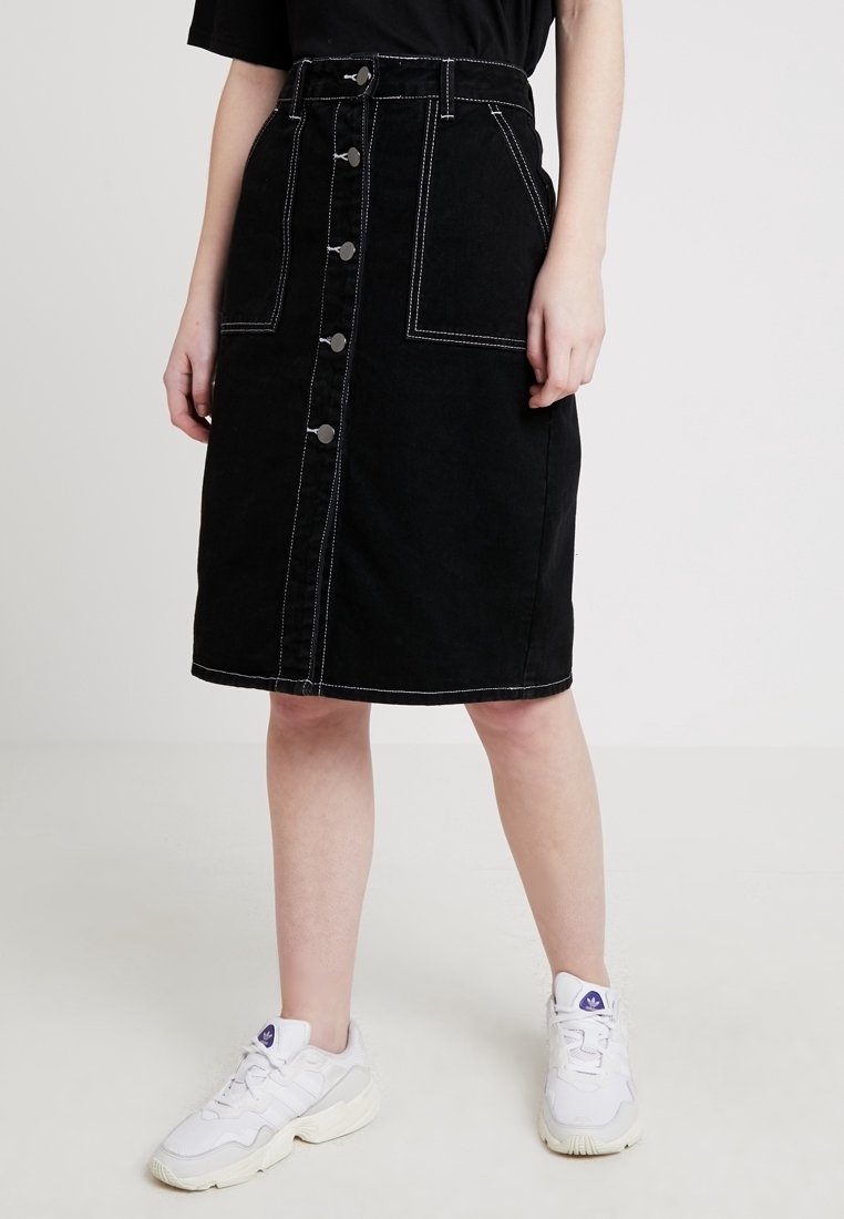 Falda midi de mezclilla negra con cierre de botones en el frente, dos grandes bolsillos, acentos de costura blanca y una silueta recta. Combinada con zapatillas blancas.