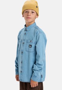 Niño con gorro de punto amarillo, camisa de mezclilla azul claro con botones y bolsillos de parche, y pantalones de pana marrones sobre un fondo blanco.