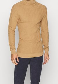 Beiger Strickpullover mit strukturiertem Rautenmuster, Rollkragen sowie gerippten Bündchen und Saum. Getragen über dunkelblauer Hose.