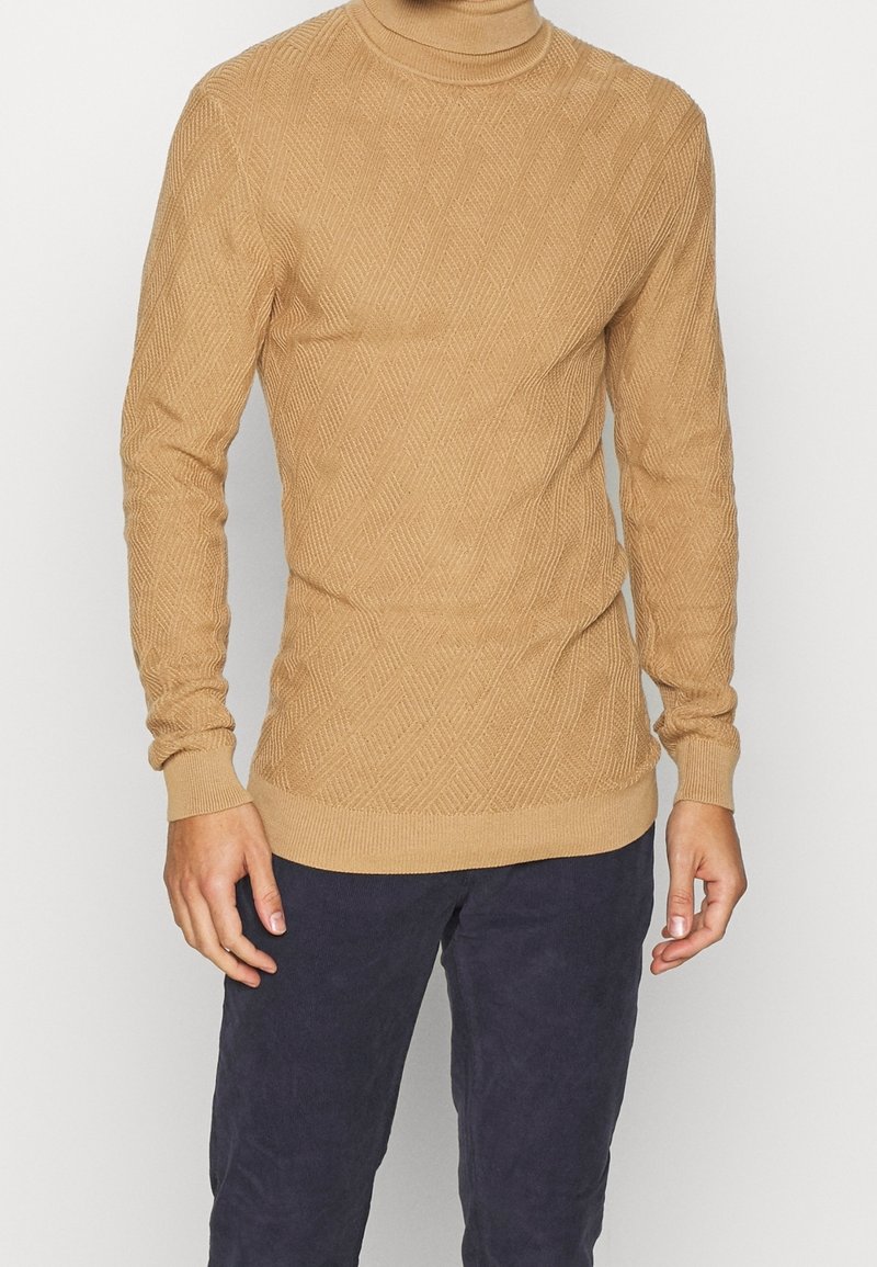 Beiger Strickpullover mit strukturiertem Rautenmuster, Rollkragen sowie gerippten Bündchen und Saum. Getragen über dunkelblauer Hose.