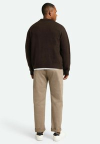 Suéter marrón de punto con cuello acanalado, llevado sobre una camisa blanca, combinado con pantalones beige y zapatillas blancas. Diseño simple y corte relajado.