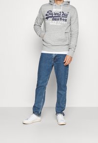 Grå huvtröja med en ficka fram och vita dragsnören, med en marinblå logotext, tillsammans med blå denimjeans och vita sneakers.