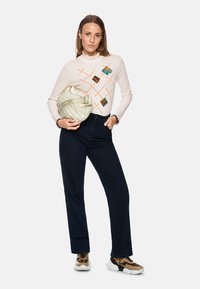 Weißes, langärmliges Oberteil mit Rüschenausschnitt und bunten geometrischen Mustern. Dunkelblaue Jeans und eine strukturierte, beige gesteppte Tasche. Sneakers im Tiermotiv.