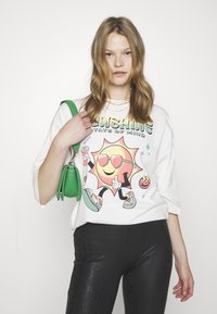T-shirt blanche oversize avec un graphique de soleil coloré et du texte, associé à des leggings noirs texturés et un petit sac vert.