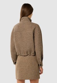 Brun houndstooth-mønstret beklædning med høj krave, lange ærmer og elastisk talje. Har en afslappet pasform og knappede ærmer.