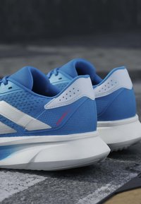 Paire de chaussures de course athlétiques bleues et blanches avec l'étiquette « DURAMO 9.2 », placées sur une surface en asphalte.