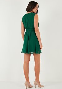 Groene chiffon jurk met een getailleerde taille en een uitlopende rok. Heeft een mouwloos ontwerp en een gladde textuur. Gekenmerkt door naaktkleurige hakken.