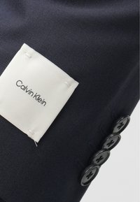 Ναυτικό ύφασμα με ετικέτα Calvin Klein σε λευκό. Διαθέτει υφασμάτινη επιφάνεια και τέσσερα μαύρα κουμπιά στην κουμπότρυπα, αναδεικνύοντας έναν κομψό σχεδιασμό.