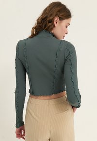 Grön ribbad långärmad crop top med volangkanter. Bakifrån visar den en figursydd design och en böjd nederkant. Matchas med krämfärgade randiga byxor.
