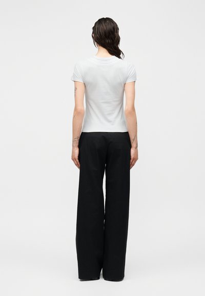 Femme aux cheveux bruns ondulés portant un haut blanc ajusté à manches courtes et un pantalon noir à larges jambes, debout avec les bras détendus le long du corps, vue de dos.