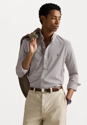 Striped button-down shirt in red and white, paired with beige trousers. Brown leather belt and wristwatch. Jacket over shoulder, clean design.