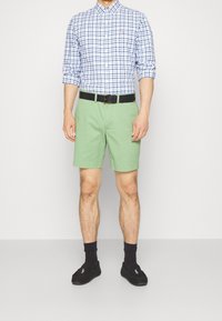 Ljusgröna shorts med rak skärning och uppvikt fåll, tillsammans med en blå och vit rutig skjorta med knappar. Svart bälte och svarta loafers.