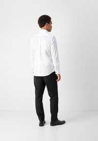 Fehér hosszúujjú ing hátvágással, fekete nadrággal és fekete cipővel párosítva, sima anyaggal és klasszikus gallér kialakítással.