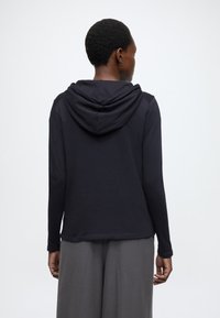 Čierna mikina s dlhými rukávmi, voľného strihu a mäkkej textúry, vybavená splývavou kapucňou a hladkými švami na ramenách.