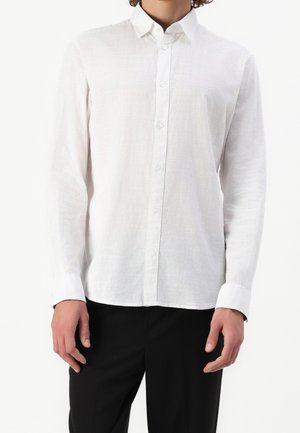 Homme portant une chemise blanche à manches longues avec col et un pantalon noir, debout devant un fond blanc uni.