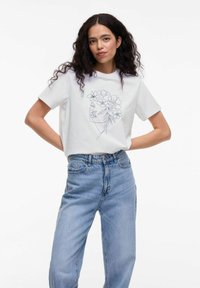 Hvid bomulds t-shirt med korte ærmer, der viser en sort linjetegning af et ansigt prydet med blomster; parret med højhalsede blå jeans.