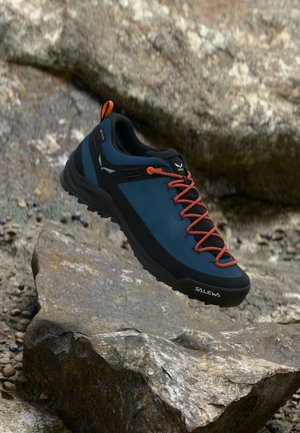 Blauw en zwarte wandelschoen met oranje veters en accenten. Heeft een robuuste zool en neuskap, ontworpen voor gebruik buitenshuis. Logo zichtbaar aan de zijkant.