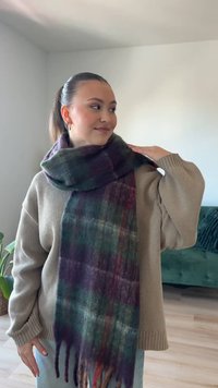 O eșarfă din lână cu un model în carouri în nuanțe de verde închis, mov și roșu, cu detalii cu franjuri la capete, așezată peste un pulover bej.