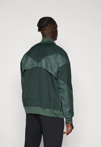 Green sports jacket with a textured, patterned back panel. Features a high collar and ribbed cuffs and hem. Durable fabric.