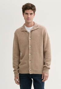 Beige gestrickte Strickjacke mit Schalkragen, sieben Kn�öpfen und gerippten Bündchen. Sie hat eine gesprenkelte Textur und einen lässigen Schnitt, kombiniert mit dunklen Jeans.