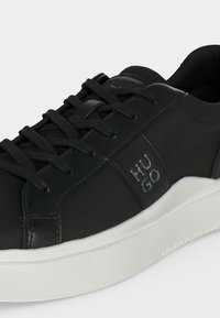 Baskets basses noires avec lacets noirs, dessus texturé, logo « HUGO » sur le côté, et semelle épaisse en caoutchouc blanc.