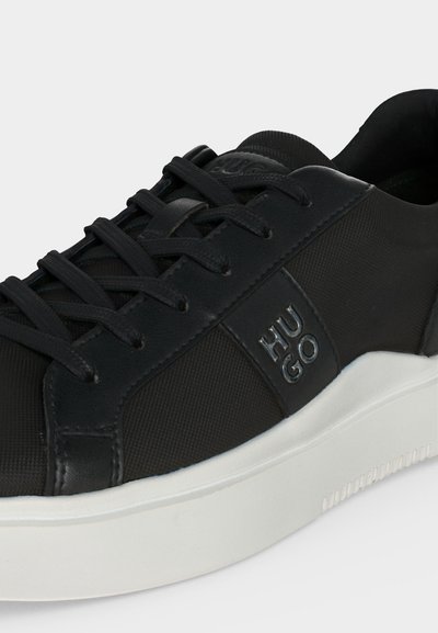 Baskets basses noires avec lacets noirs, dessus texturé, logo « HUGO » sur le côté, et semelle épaisse en caoutchouc blanc.