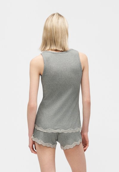 Rövid szőke hajú nő, szürke ujjatlan felsőt és hozzá illő rövidnadrágot visel, fehér csipkés díszítéssel, hátulról látható.
