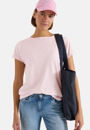 Femme portant une casquette rose, un t-shirt rayé rose clair, un jean bleu, portant un sac fourre-tout noir sur l'épaule devant un fond blanc.