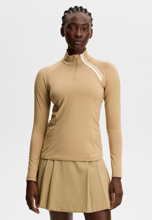 Kvinde med fletninger iført en beige langærmet lynlåstop med hvide striber og et matchende plisseret skørt mod en ensfarvet baggrund.