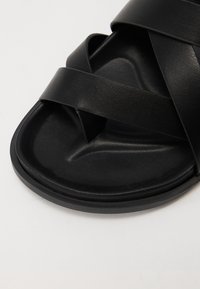 Black leather sandal with crisscross straps, flat sole, and textured footbed. Features a minimalist design and smooth finish.
