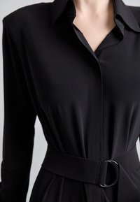 Robe noire à manches longues avec col et large ceinture assortie ornée d'une boucle ronde en métal, cintrée à la taille.