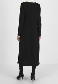 Vestido negro de manga larga con silueta recta, cierre de cremallera en la parte posterior y escote modesto, combinado con botas de tacón negras hasta la rodilla.
