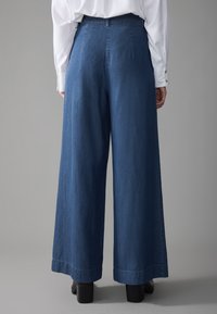 Pantalones de mezclilla de pierna ancha en azul oscuro, con una cintura alta, una textura sutil y un dobladillo limpio. Combinados con zapatos negros de tacón bloque.