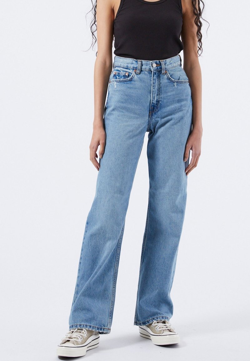 Personne portant un jean bleu clair taille haute à jambes larges, des baskets beiges, et un haut noir sans manches, debout devant un fond uni.