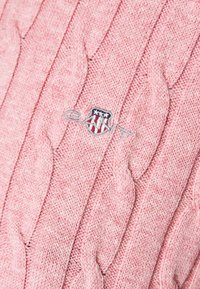 Różowy sweter w sploty z fakturowanymi warkoczami. Haftowane logo w kolorze niebieskim, białym i czerwonym na środku. Miękka tkanina o ciepłym wyglądzie.