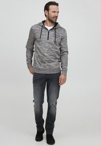 Grå hettegenser med et teksturert mønster, frontglidelås og snorer. Kombinert med mørke jeans og svarte sko, vist mot en ensfarget bakgrunn.