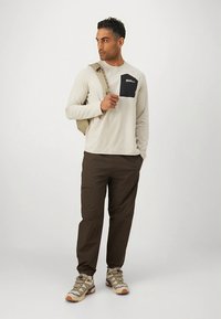 Beiges Langarmshirt mit einer dunkelblauen Brusttasche, kombiniert mit braunen Cargohosen und grauen Wanderschuhen. Beige Rucksack mit einem Gurt.