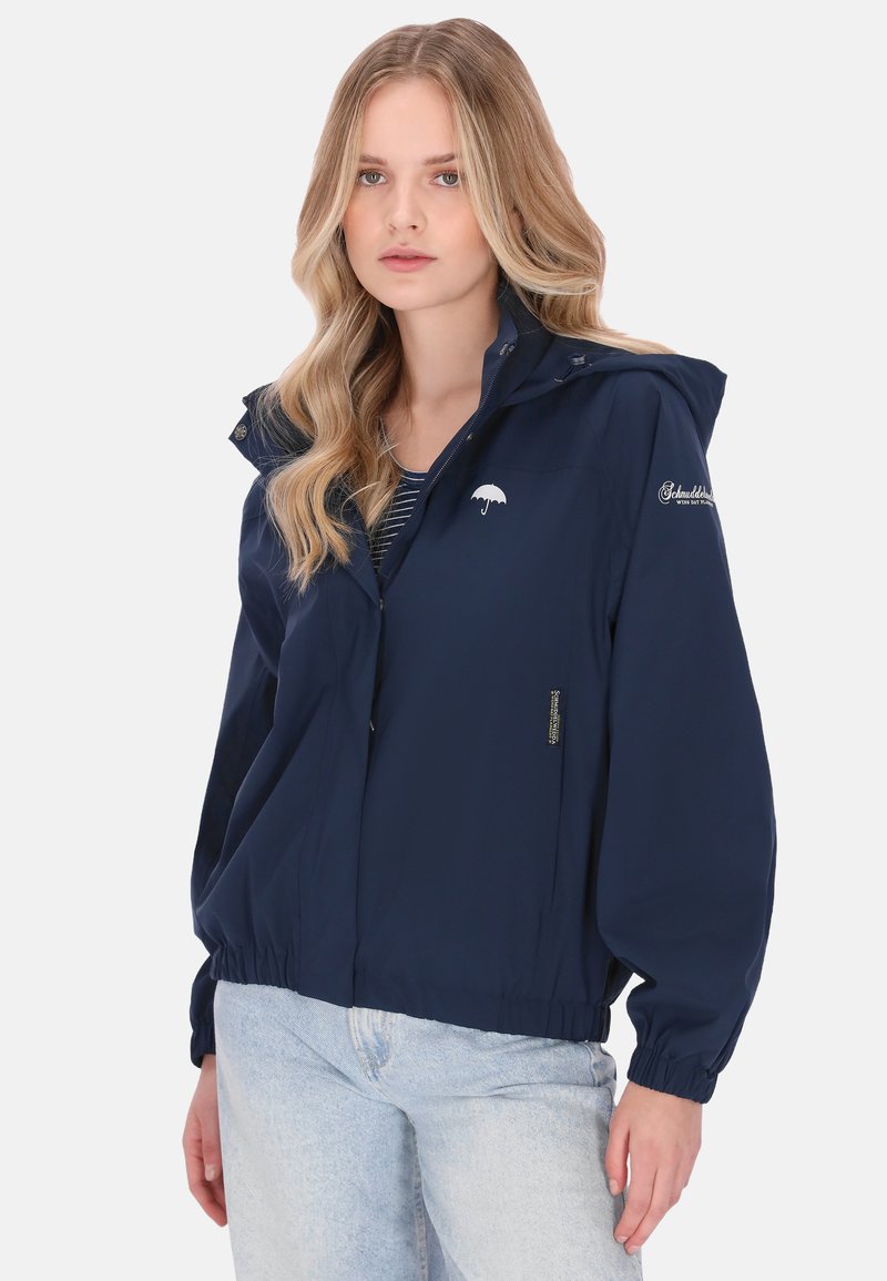 Granatowa wodoodporna kurtka z kapturem, elastycznymi mankietami i dolnym obszyciem. Posiada minimalistyczne logo parasola. Do tego jasnoniebieskie dżinsy.