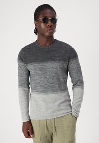 Cămașă tricotată cu mânecă lungă în degradé de la gri închis la gri deschis, guler rotund, detalii ribbate, purtată cu pantaloni cargo și ochelari de soare.
